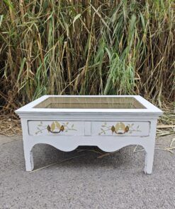 Mesa de centro shabby chic con tapa de cristal, detalles decorativos y acabado blanco envejecido.